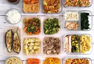 Batch cooking containers filled with healthy prepared meals including vegetables, rice, pasta, and curry dishes on a white wooden table.